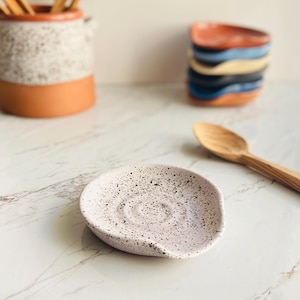 May include: A speckled, pale pink ceramic spoon rest with a curved design. The rest has a small indentation for holding a spoon. A wooden spoon and other ceramic kitchenware are in the background.