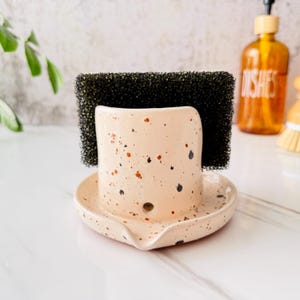 Può includere: Un porta spugna in ceramica maculata color pesca chiaro con macchie nere e marroni. Il supporto ha un vassoio integrato e contiene una spugna nera. Sullo sfondo c'è una bottiglia di detersivo per piatti.