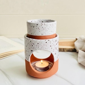 May include: A ceramic essential oil diffuser with a speckled white and terracotta design. The diffuser has a circular opening to hold a tea light candle, which is lit. The top section is designed to hold essential oils.