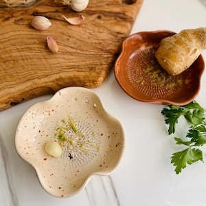 May include: A beige ceramic garlic grater with a flower-like shape, speckled with red and black dots. A clove of garlic sits on the grater. A brown ceramic ginger grater is also visible, along with a wooden cutting board and fresh parsley.