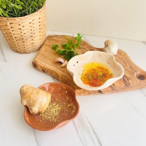 May include: A ceramic ginger grater and a small, speckled, wavy-edged bowl with oil and spices sit on a wooden cutting board. Fresh ginger, garlic, and parsley are also present, suggesting a culinary theme.