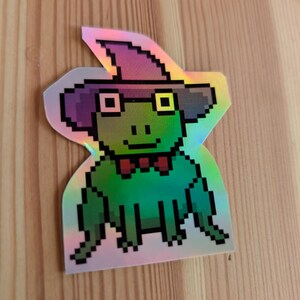 Peut inclure: Autocollant holographique représentant une grenouille verte pixélisée portant un chapeau de sorcière violet, des lunettes carrées et un nœud papillon rouge. La grenouille sourit. Fond en bois.