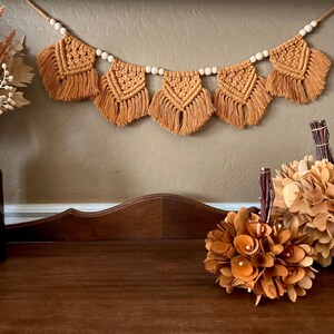 May include: A brown wooden table with a tan macrame garland hanging on the wall above it. Two decorative pumpkins made of wood are on the table. The pumpkins are brown and have a flower-like design.