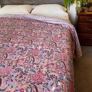 May include: A pink and blue floral patterned quilt with a white border. The quilt is on a bed with white pillows and a wooden headboard.