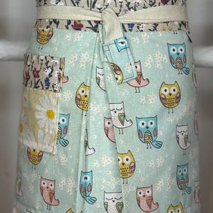 May include: A light blue apron with an owl pattern. The apron has a floral waistband and pocket, with a daisy pattern on the pocket. It ties at the waist with a cream-coloured tie.