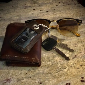 May include: A brown leather wallet with a key fob attached. The key fob has a small black remote with the words "LOCK" and "UNLOCK" on it. The key fob is also attached to a set of keys and a pair of brown and gold aviator sunglasses.