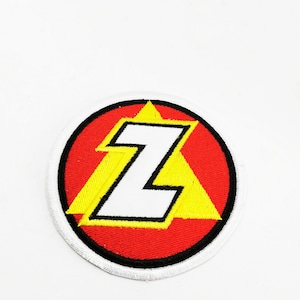 Puede incluir: Parche circular con una audaz letra "Z" blanca con contorno negro, sobre un triángulo amarillo dentro de un círculo rojo. El parche tiene un borde blanco. Probablemente un logotipo o emblema.