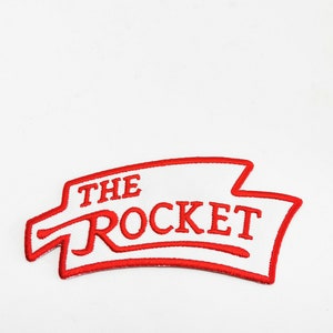 以下が含まれることがあります： 白い背景に赤い文字で「THE ROCKET」と刺繍されたパッチ。赤い縁取りがあり、ユニークな形をしており、ジャケット、バッグ、その他の布製品に適しています。