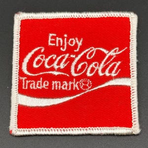 Peut inclure: Un écusson carré brodé avec un fond rouge vif et une bordure blanche cousue. L'écusson présente le texte brodé en blanc Enjoy Coca-Cola et Trade mark, ainsi qu'une ligne courbe blanche sur la partie inférieure.