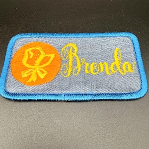 May include: A rectangular embroidered patch with a light blue denim-textured background and a bright blue stitched border. On the left, an orange circular emblem features a yellow stylized bird graphic. The name Brenda is embroidered in yellow script.