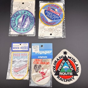 Puede incluir: Cinco parches bordados de recuerdo con temas de Pikes Peak y Colorado. Los diseños incluyen formas circulares, triangulares y de escudo con textos como I MADE IT SUMMIT PIKES PEAK 14,110 FT, SKI COLORADO y COLORADO MIDLAND PIKES PEAK ROUTE RAILWAY.