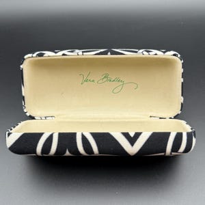 Könnte beinhalten: Geöffnete Vera Bradley Brillenetui. Das Etui ist schwarz-weiß mit einem geometrischen Muster. Die Innenseite ist beige mit dem Vera Bradley-Logo in grüner Schrift.