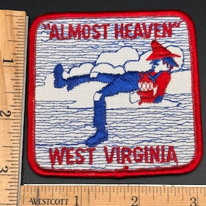 May include: A square embroidered patch with a red border, featuring a blue figure in a red hat holding a jug, set against a white background with blue wavy lines. The text ALMOST HEAVEN and WEST VIRGINIA is in red. A ruler shows it is 3 inches wide.