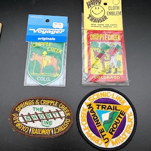 May include: Four embroidered patches featuring Cripple Creek, Colorado themes. Two are in plastic packaging with header cards. One is oval with a railway graphic, and one is circular with a yellow triangle and purple border design.