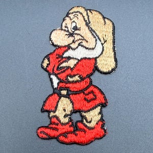 May include: An embroidered patch featuring a cartoon character with a grumpy expression, wearing a tan hat, a red tunic with a black belt, and red shoes. The character has a white beard and is depicted with arms crossed in a standing position.