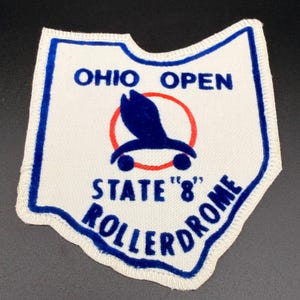 May include: A white fabric patch shaped like the state of Ohio with a blue border. It features the text OHIO OPEN, STATE '8, and ROLLERDROME in blue, along with a central graphic of a dark blue bird silhouette inside a thin red circle.