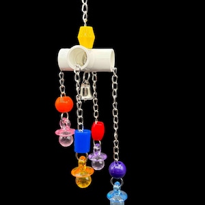 Puede incluir: Un colorido juguete para pájaros con una base de tubo de PVC blanco, cadenas colgantes y varias formas de plástico. Incluye una pequeña campana y una forma geométrica amarilla en la parte superior. El juguete presenta detalles en naranja, rosa, azul, rojo, morado y amarillo.