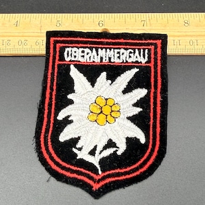 Puede incluir: Un parche bordado negro que presenta una flor de edelweiss blanca con un centro amarillo y el texto OBERAMMERGAU en blanco. El parche tiene forma de escudo con un borde rojo y está colocado debajo de una regla de madera de unos 9 cm.