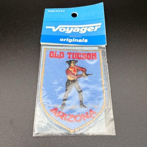 Op de afbeelding: Een schildvormige geborduurde patch in een doorzichtige plastic hoes met een blauwe koptekst met de tekst Voyager originals. De patch heeft een lichtblauwe achtergrond, een figuur in een rood geruit overhemd en donkere broek, en rode tekst Old Tucson Arizona.