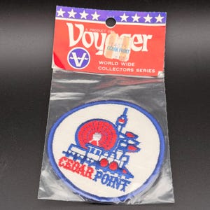 May include: A round embroidered patch featuring a red Ferris wheel and a blue train engine with the text Cedar Point in red and blue. The patch is inside a clear plastic bag with a red and white header card labeled Voyager World Wide Collectors Series.