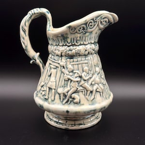 May include: A ceramic pitcher featuring a cream-colored base with blue-green glaze accents. The exterior displays a raised relief scene of figures in a tavern setting, a decorative border near the rim, and a curved handle with a textured finish.