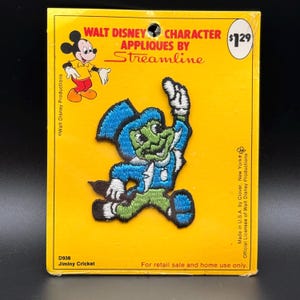 May include: A Walt Disney Character applique by Streamline featuring an embroidered Jiminy Cricket patch on a yellow backing card. The patch depicts the character in a blue top hat and jacket with green trousers, white gloves, and black shoes.