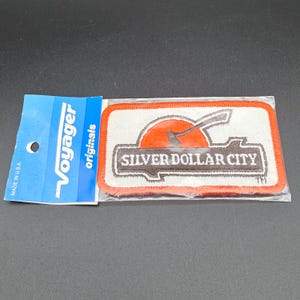 May include: A rectangular embroidered patch featuring the text Silver Dollar City in white letters on a brown log graphic, set against an orange sun and white background with an orange border, attached to a blue Voyager Originals cardboard header.