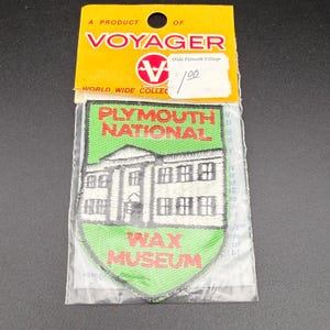 May include: A shield-shaped embroidered patch in a clear plastic bag with a yellow header card labeled VOYAGER. The patch features a green background, a white building illustration, and red text reading PLYMOUTH NATIONAL WAX MUSEUM.