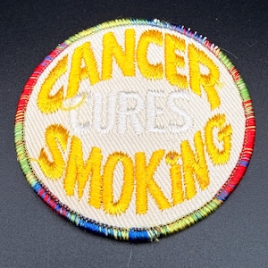 May include: A circular embroidered patch with a beige background and a multicolored border. The text CANCER CURES SMOKING is stitched in bright yellow thread, with the word CURES appearing in a lighter, subtle off-white thread color.