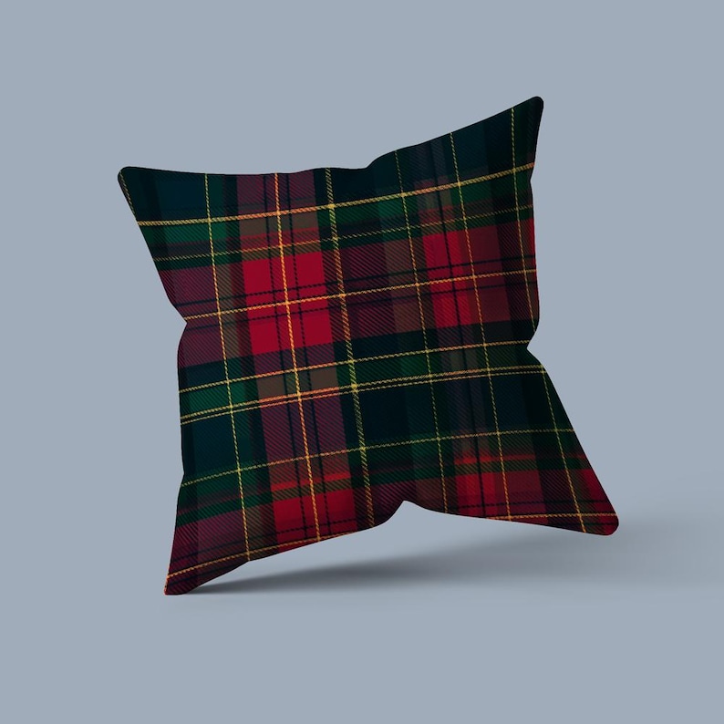 May include: Square decorative pillow with a classic tartan plaid pattern in shades of red, green, navy, and yellow. The pillow has a soft, textured appearance and is likely made of fabric. The design is symmetrical and the pillow is displayed on a light blue background.