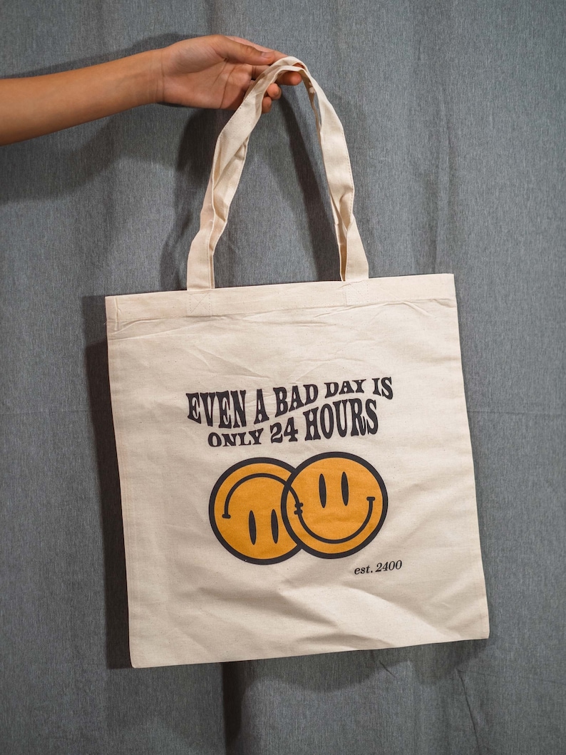 May include: Cream-colored tote bag with the text "EVEN A BAD DAY IS ONLY 24 HOURS" and two overlapping yellow smiley faces. The bag has two cream-colored handles and the text "est. 2400".