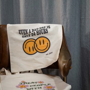 May include: Off-white canvas tote bags with positive messages and smiley face graphics. One bag reads "EVEN A BAD DAY IS ONLY 24 HOURS" with two overlapping smiley faces. Another bag says "TREAT PEOPLE WITH KINDNESS" with flower accents.