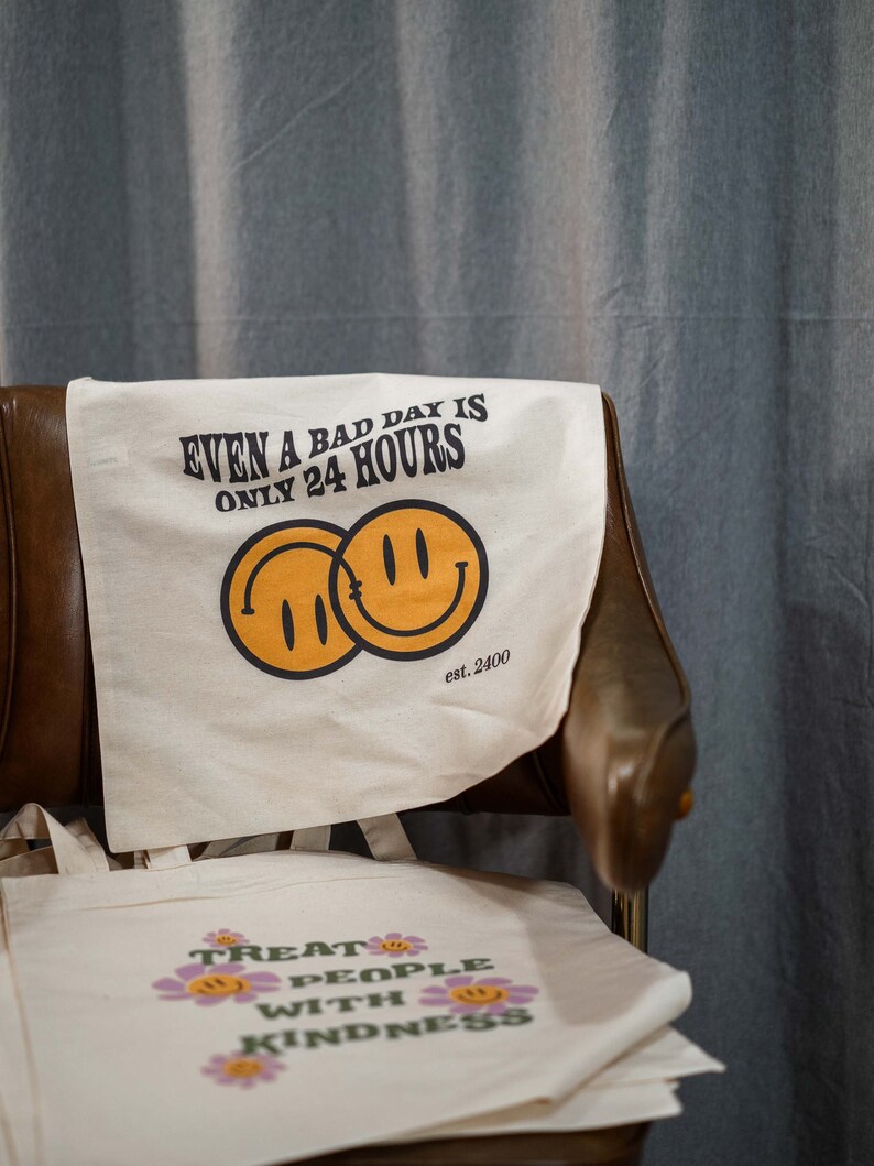 May include: Off-white canvas tote bags with positive messages and smiley face graphics. One bag reads "EVEN A BAD DAY IS ONLY 24 HOURS" with two overlapping smiley faces. Another bag says "TREAT PEOPLE WITH KINDNESS" with flower accents.