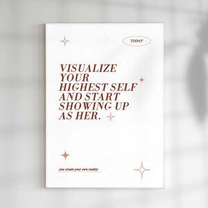 May include: A white framed art print with the text "VISUALIZE YOUR HIGHEST SELF AND START SHOWING UP AS HER." in a rust-colored font. The word "TODAY" is above the text, and "you create your own reality" is below. Decorative starbursts are also included.