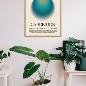 May include: A framed Capricorn zodiac sign art print. The artwork features a teal and orange sphere on a cream background, with the word "CAPRICORN" and related traits below. The print is displayed in a room with plants.