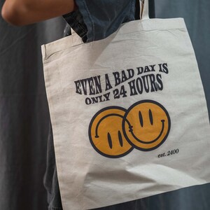 May include: Beige tote bag with the text "EVEN A BAD DAY IS ONLY 24 HOURS" in dark brown. Two overlapping yellow smiley faces are below the text, with "est. 2400" below them. The bag is held by a person.