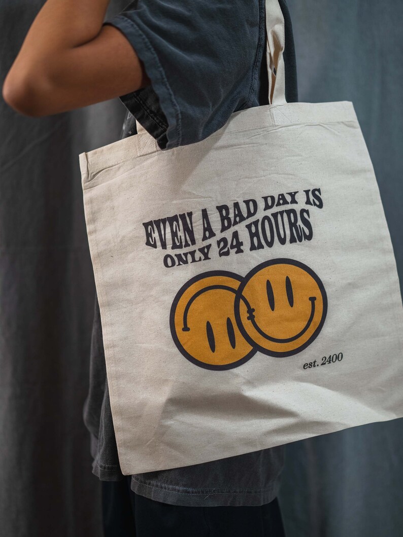 May include: Beige tote bag with the text "EVEN A BAD DAY IS ONLY 24 HOURS" in dark brown. Two overlapping yellow smiley faces are below the text, with "est. 2400" below them. The bag is held by a person.