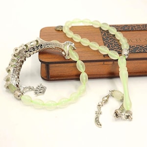 May include: A light green prayer bead set and a bracelet with silver-colored metal accents. The bracelet has the text "MULTI BRACELET" and a small crown charm. The beads are oval-shaped and rest on a wooden box.