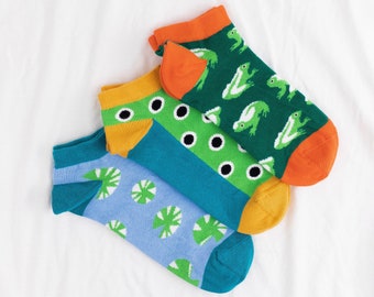 May include: Three pairs of ankle socks with different patterns. One pair is blue with green lily pads, one pair is orange with green and white frogs, and one pair is teal with green circles and black dots.