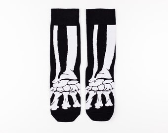 May include: A pair of black and white socks with a skeleton foot design. The socks feature a black background with a white bone graphic of the foot and ankle. The socks are displayed on a white background.
