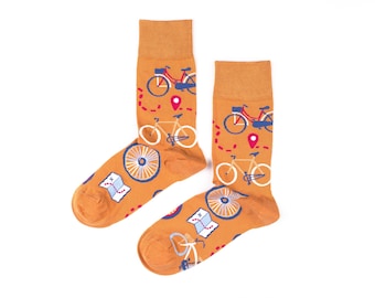 May include: A pair of orange socks with a bicycle theme. The socks feature illustrations of bicycles, maps, and wheel designs in white, blue, and red. The socks are on a white background.