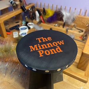 Könnte beinhalten: Ein schwarzer Hocker mit dem Text "The Minnow Pond" in Orange. Der Hocker hat eine runde Oberseite mit weißen Akzenten und einem dunklen Sockel. Der Hintergrund zeigt Werkzeuge und Materialien für das Handwerk.