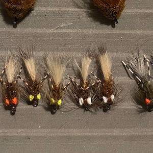 May include: Assortment of fly fishing lures with different colored accents. The lures have brown, black, and white feathers and are arranged in a row.