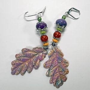 May include: A pair of dangle earrings featuring iridescent purple and gold oak leaf charms. Each earring has a silver-tone leverback hook and a vertical arrangement of beads in green, purple, red, orange, and dark gray with silver-tone bead caps.