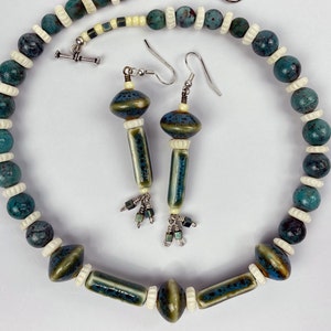 May include: A jewelry set consisting of a necklace and matching drop earrings. The necklace features round teal speckled beads, white ribbed spacers, and elongated ceramic tube beads. The earrings mirror this design with silver hooks.