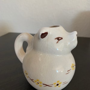Op de afbeelding: Een kleine, witte keramische creamer in de vorm van een berenkop. Het heeft bruine accenten voor de ogen en oren, en een decoratief bloemmotief in geel en bruin. De creamer heeft een handvat en een schenktuit.