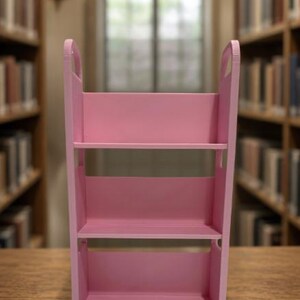 May include: A pink three-tiered rolling utility cart with open shelves and side handles. The cart features a rectangular frame with four small wheels at the base, providing a storage solution with three distinct levels for organizing various items.