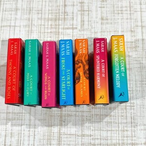 May include: Seven miniature books by Sarah J. Maas arranged in a row. The spines are colored red, green, pink, blue, orange, magenta, and yellow-to-teal gradient, featuring titles like A Court of Thorns and Roses and A Court of Silver Flames.