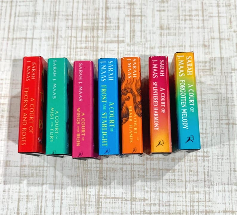 May include: Seven miniature books by Sarah J. Maas arranged in a row. The spines are colored red, green, pink, blue, orange, magenta, and yellow-to-teal gradient, featuring titles like A Court of Thorns and Roses and A Court of Silver Flames.