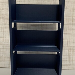 May include: A dark navy blue three-tiered rolling cart featuring a rectangular frame with two vertical side panels, integrated handles at the top, and four small wheels at the base. The unit has three open shelves with solid back panels.